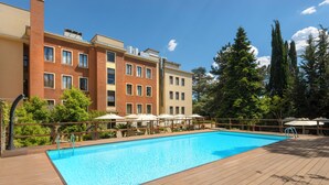 A swimming pool with a wooden deck, surrounded by a multi-story building with balconies and trees. - Occidental Aurelia (Rome)