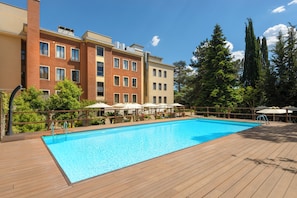 A swimming pool with a wooden deck, surrounded by a multi-story building with balconies and trees. - Occidental Aurelia (Rome)
