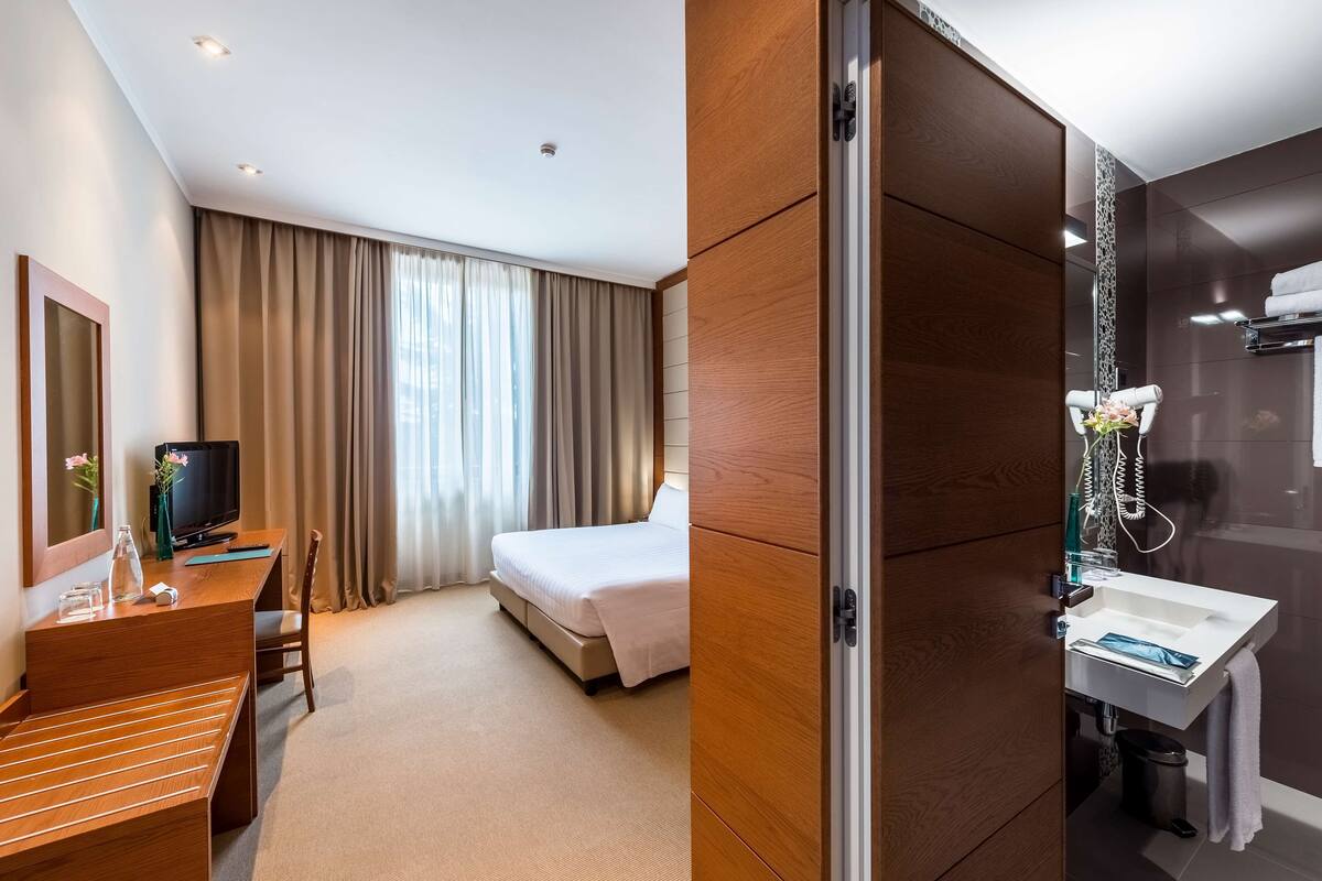 Habitación de hotel con cama, escritorio con televisor, espejo y baño con lavamanos y ducha.