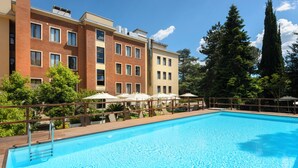 A swimming pool with a wooden deck, surrounded by a multi-story building with balconies, and trees in the background. - Occidental Aurelia (Rome)