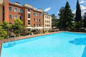 Una piscina con cubierta de madera, rodeada por un edificio de varios pisos con balcones y árboles al fondo.