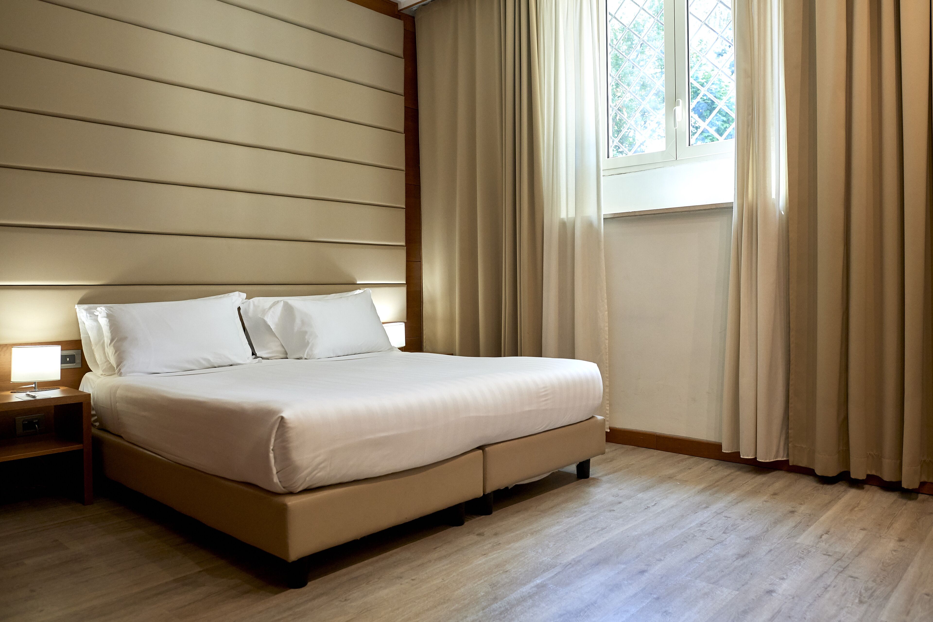 Una cama bien hecha con cabecera de madera, una mesita de noche con lámpara y una ventana con cortinas.