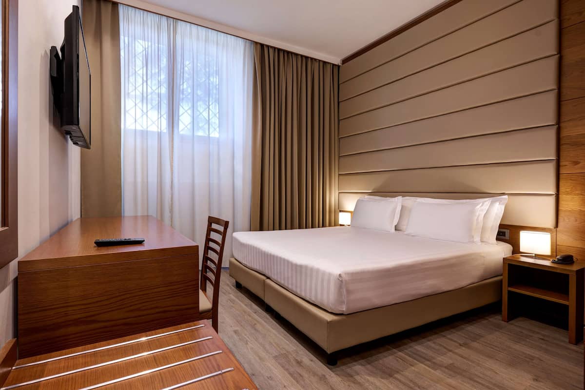 Habitación de hotel con cama, mesita de noche, silla, escritorio, televisor y cortinas.