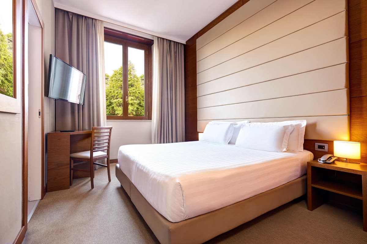 Habitación de hotel con cama, escritorio, silla, televisor y ventana con cortinas.