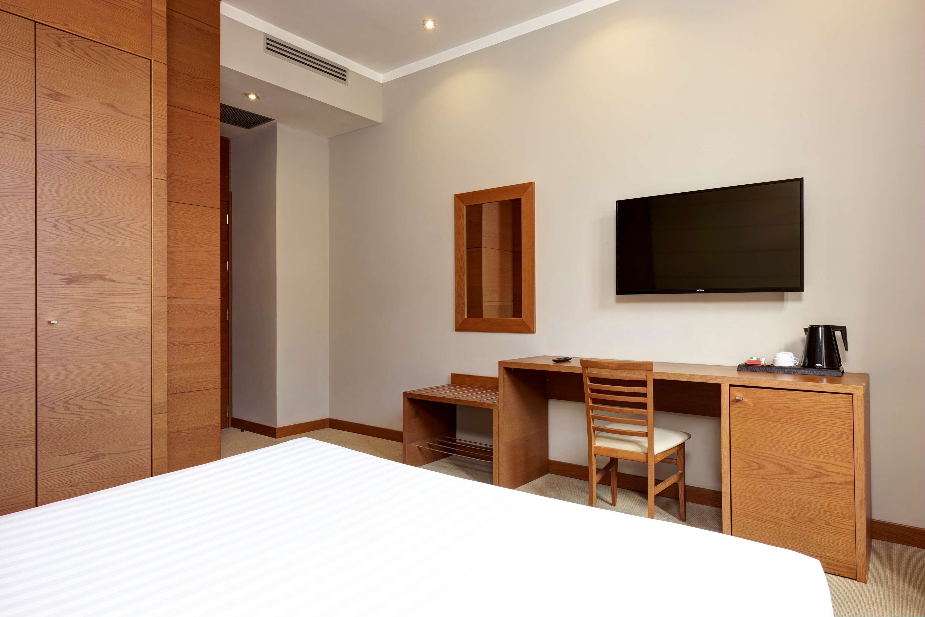 A hotel room with a bed, wooden wardrobe, desk, chair, and a flat-screen TV.
