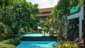 Une piscine entourée d’une végétation luxuriante, un bâtiment blanc avec un toit brun et une formation rocheuse.