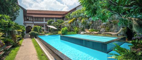 A swimming pool with a stone pathway, lush greenery, and a building with a tiled roof in the background.