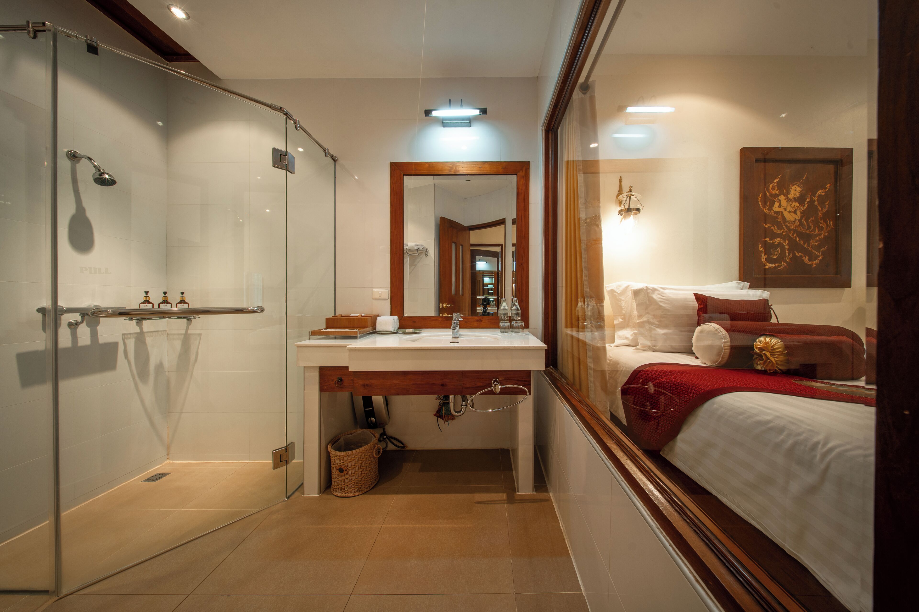 A hotel room with a glass shower enclosure, a wooden-framed mirror, a bed with red bedding, and a wall-mounted artwork.