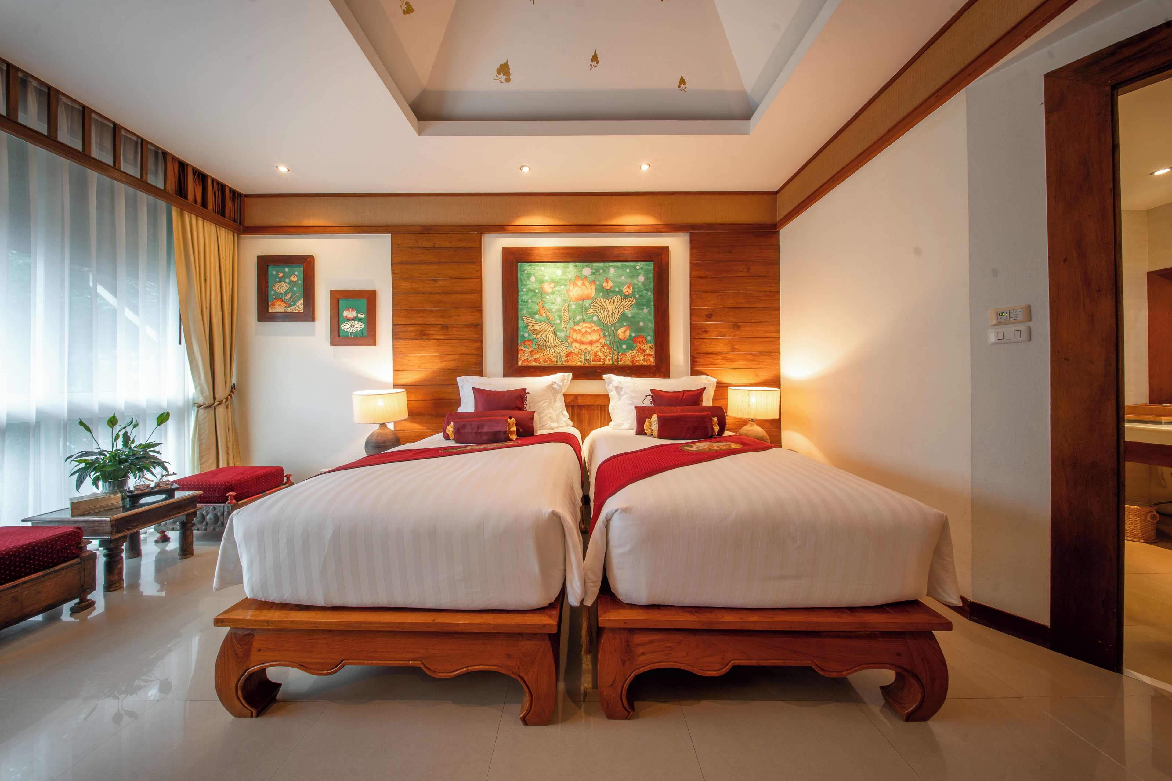 A hotel room with two beds, a wooden bench, a large window with curtains, and a painting on the wall.