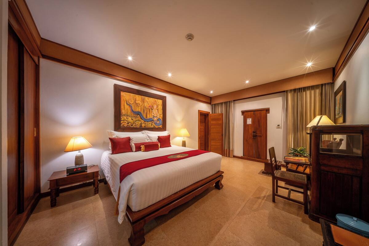 Ein Hotelzimmer mit einem großen Bett, Nachttischlampen, einem Holznachttisch, einem Schreibtisch, einem Stuhl und einem Gemälde an der Wand.