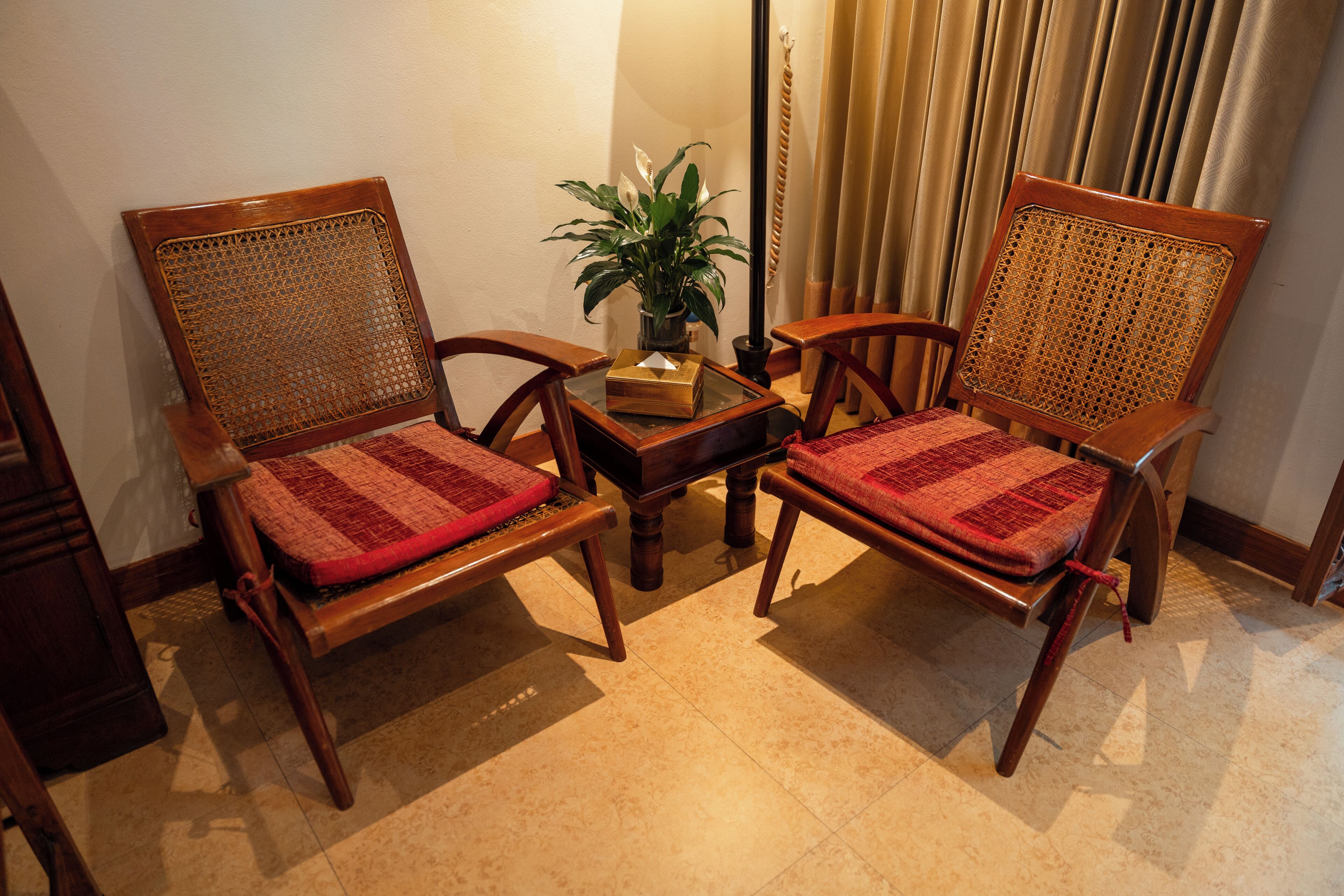 Two wooden chairs with woven backs and red cushions, a small table with a plant, and a curtain in the background.