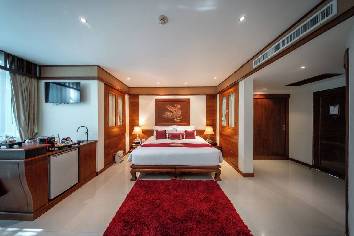 Ein Hotelzimmer mit Bett, Nachttischen, Fernseher, Schreibtisch und einem kleinen Kühlschrank, sowie einem roten Teppich.