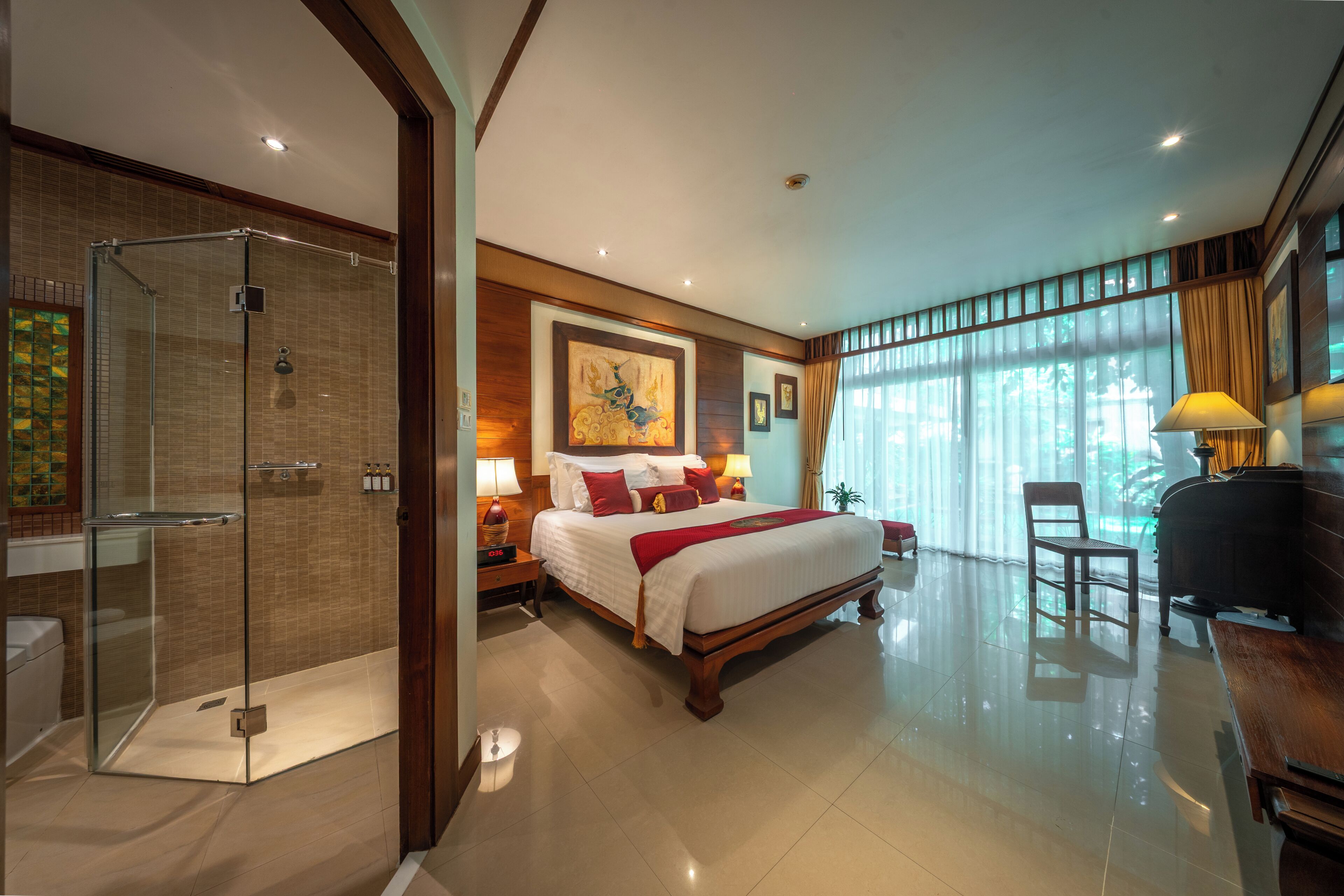 A hotel room with a glass-enclosed shower, a large bed with multiple pillows, a desk with a chair, and a large window with curtains.