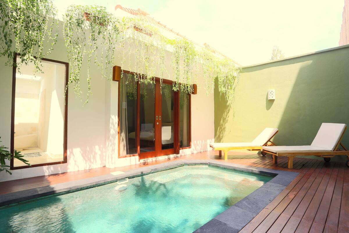 A swimming pool with a wooden deck, a glass door entrance, and a bench.