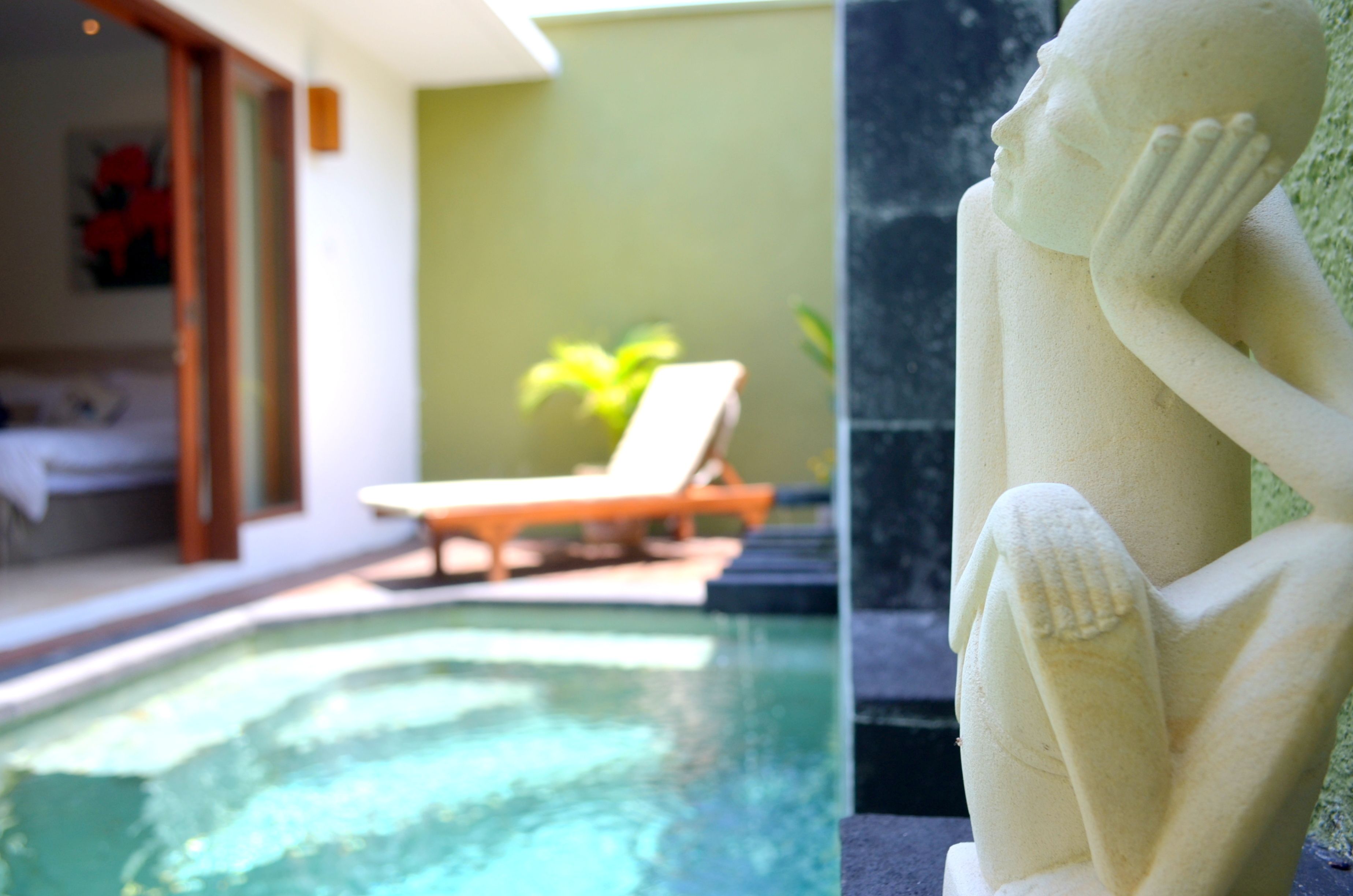 A statue of a reclining figure next to a swimming pool with a lounge chair and a plant in the background.