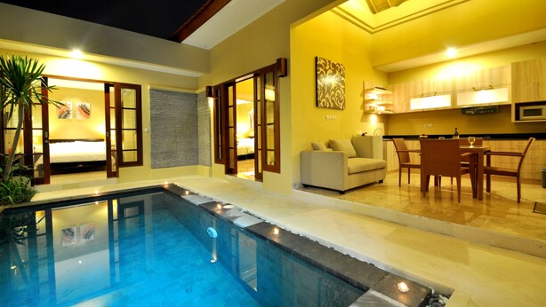 A modern indoor pool area with a clear pool, a seating area with a sofa and chairs, and a kitchen area with a dining table and chairs. - The Jas Villas (Seminyak)