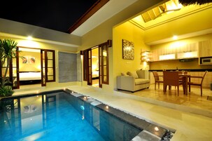 A modern indoor pool area with a clear pool, a seating area with a sofa and chairs, and a kitchen area with a dining table and chairs.