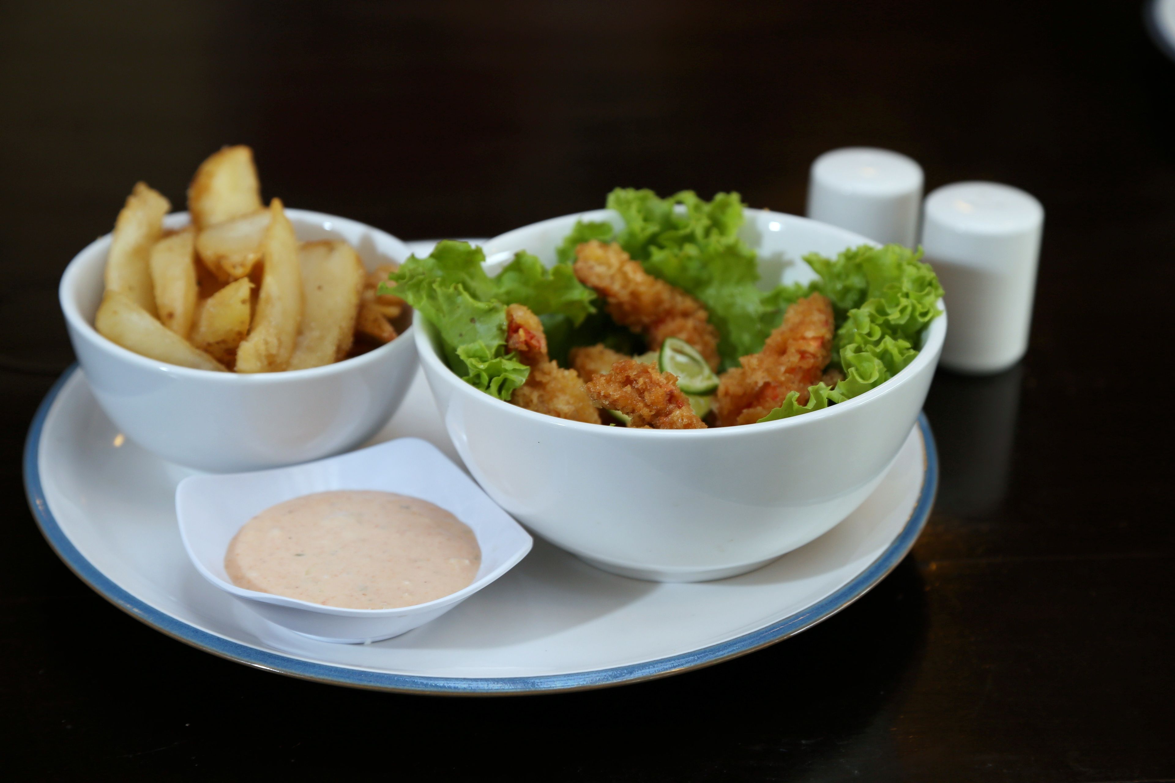 A table with two bowls of food, one containing fries and the other containing a salad with lettuce and breaded chicken pieces, and a small bowl of sauce.