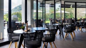 A modern outdoor dining area with round tables and black chairs, large glass windows, and a view of greenery and hills. - Handara Golf & Resort Bali (Bedugul)