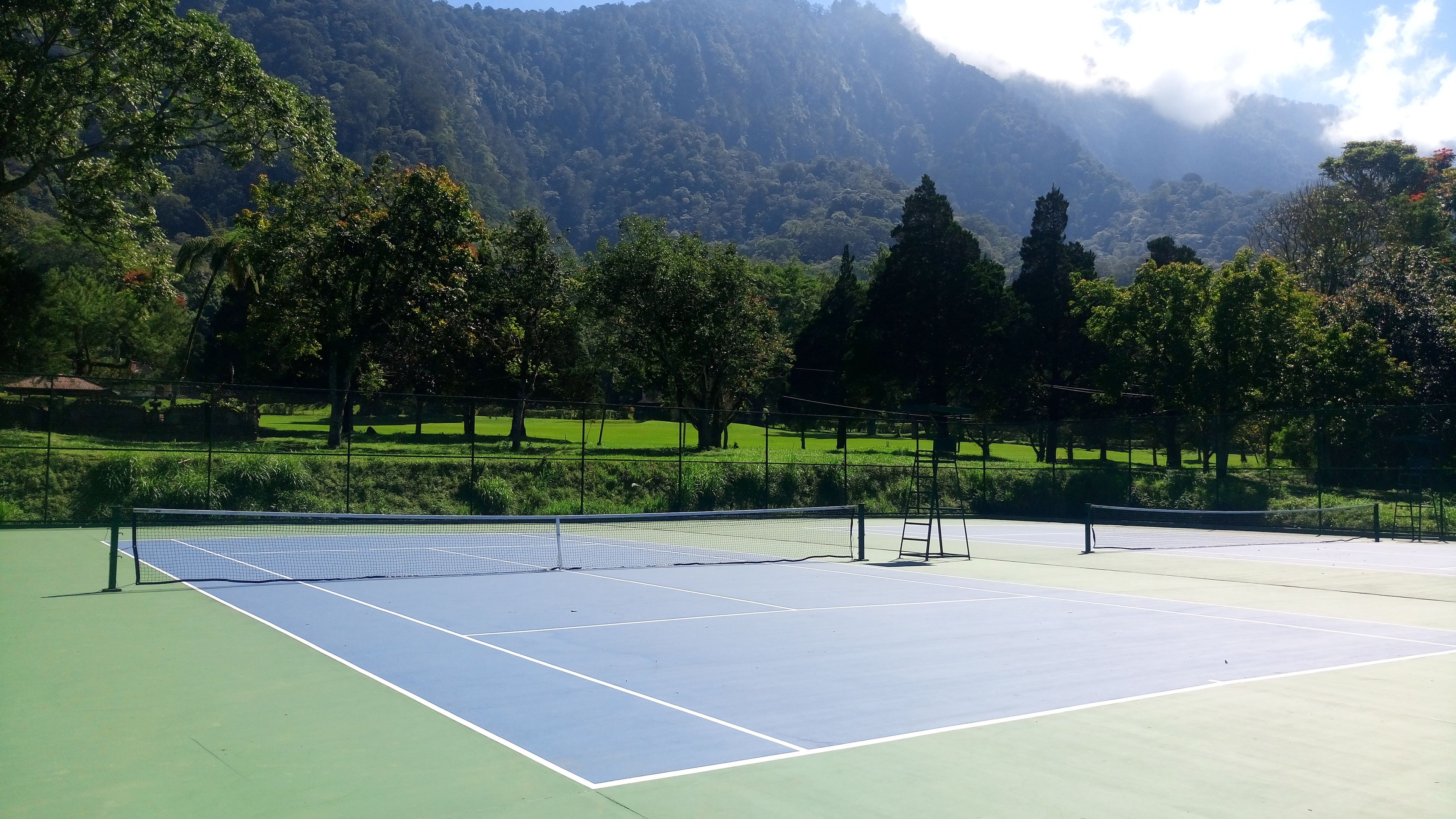 Una cancha de tenis con una red, rodeada de árboles y con montañas al fondo.
