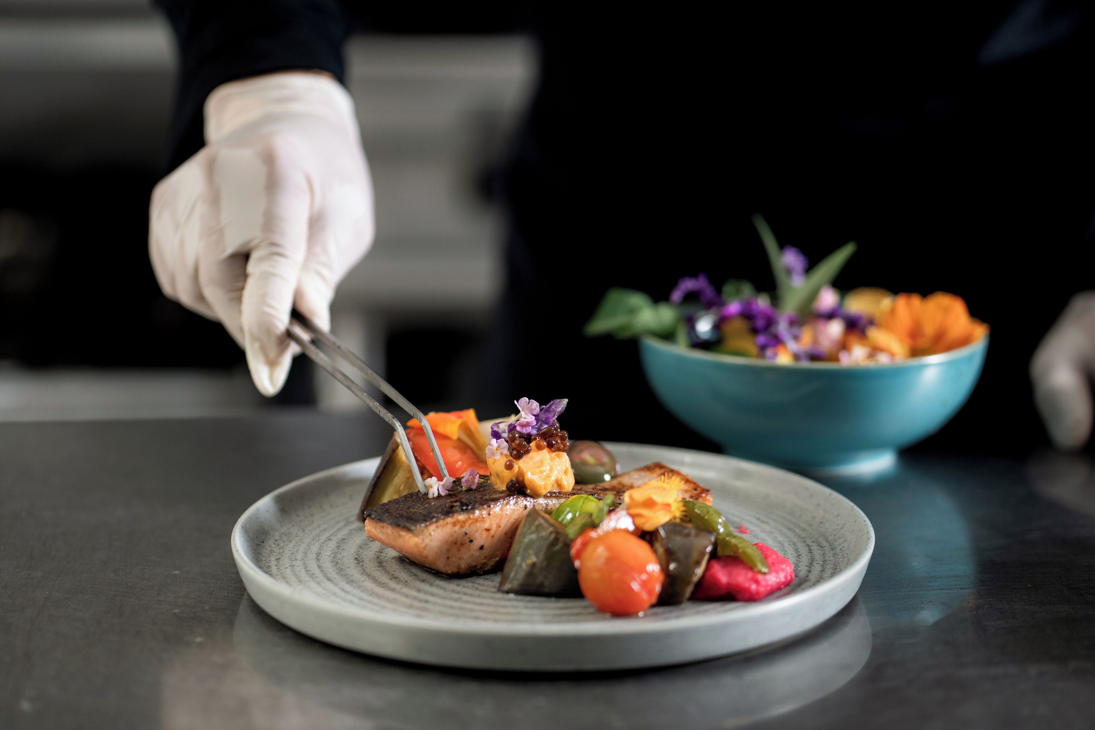 A chef's hand holding tongs over a plate of food, with a bowl of salad in the background.