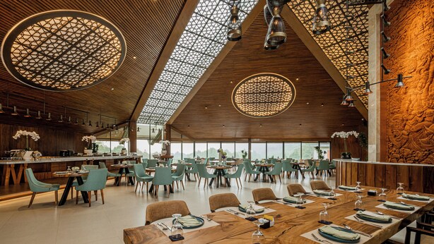 A spacious dining area with long wooden tables set for multiple guests, surrounded by modern chairs and a high ceiling with geometric patterns. - Handara Golf & Resort Bali (Bedugul)