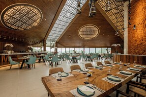 A spacious dining area with long wooden tables set for multiple guests, surrounded by modern chairs and a high ceiling with geometric patterns. - Handara Golf & Resort Bali (Bedugul)