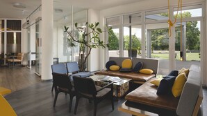 A modern living room with a sofa, chairs, and a coffee table. There is a large window with a view of the outdoors. - Sport- & Seminarhotel Glockenspitze (Altenkirchen)