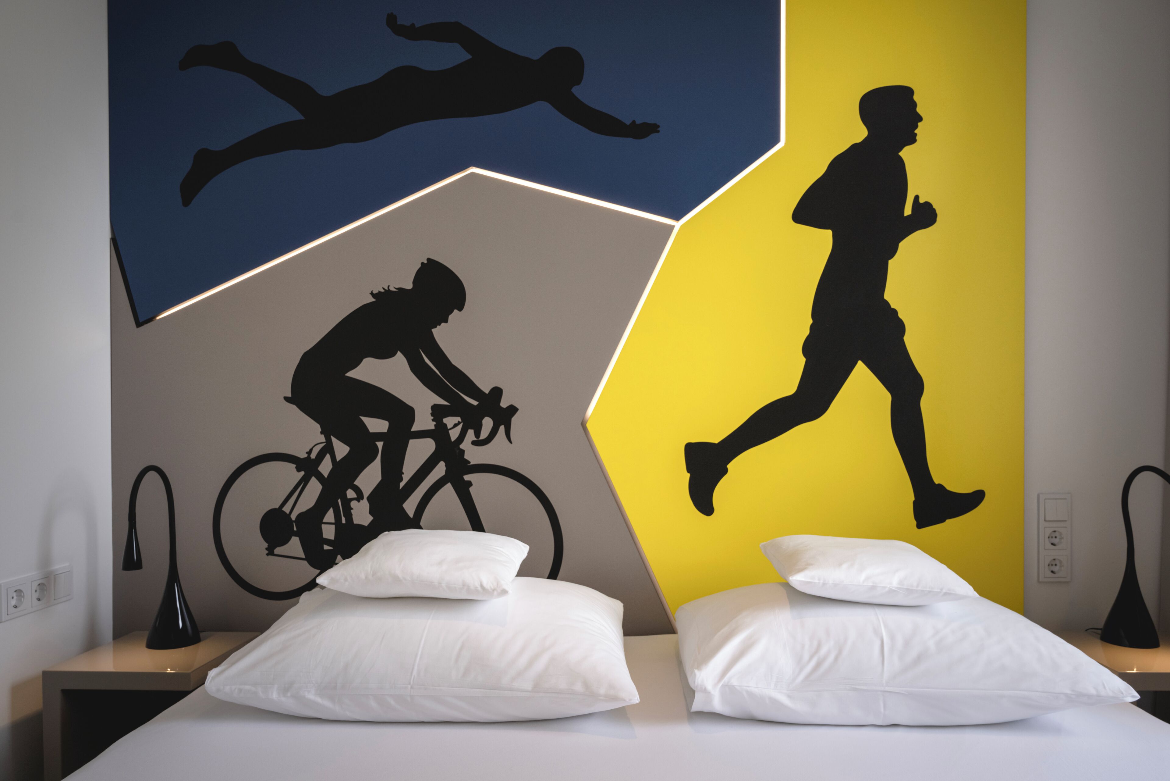 A room with a bed, pillows, a nightstand, and a wall with silhouettes of a swimmer, a cyclist, and a runner.