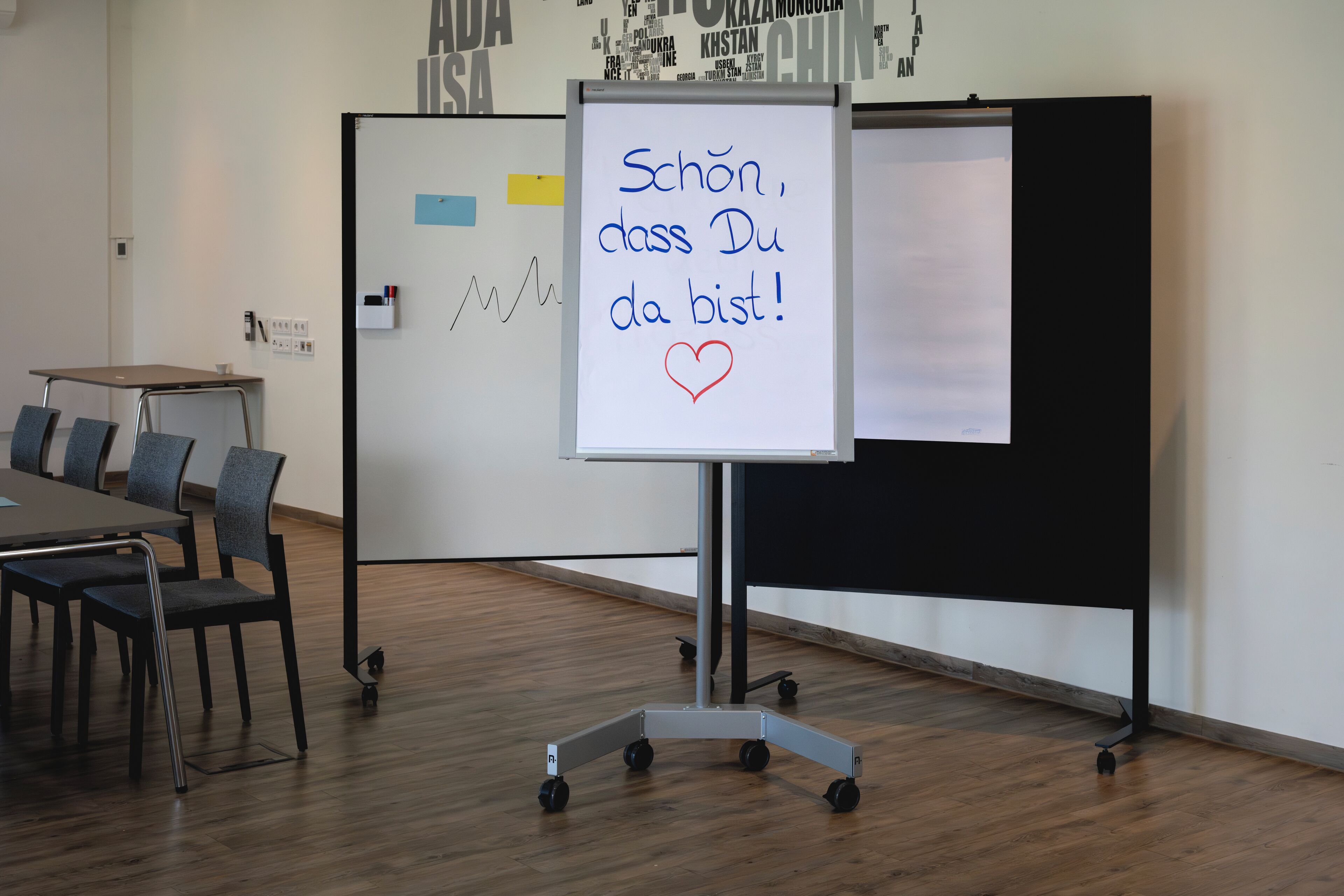 A classroom with a whiteboard displaying a message in German, a desk with chairs, and a projector screen.