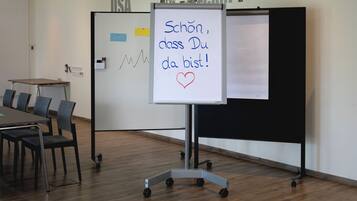 A classroom with a whiteboard displaying a message in German, a desk with chairs, and a projector screen.