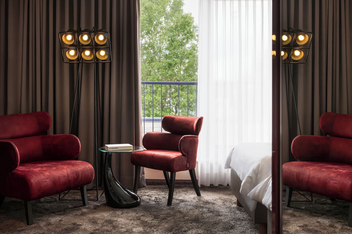 A hotel room with a large window, two red armchairs, a small table with a book, and a floor lamp.