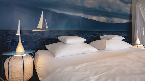 A bedroom with a bed, white pillows, a wall-mounted lamp, and a nightstand. - Sport- & Seminarhotel Glockenspitze (Altenkirchen)