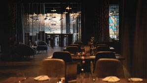 A dimly lit restaurant with tables set for dining, a bar area in the background, and large windows with a view of the cityscape. - Sport- & Seminarhotel Glockenspitze (Altenkirchen)