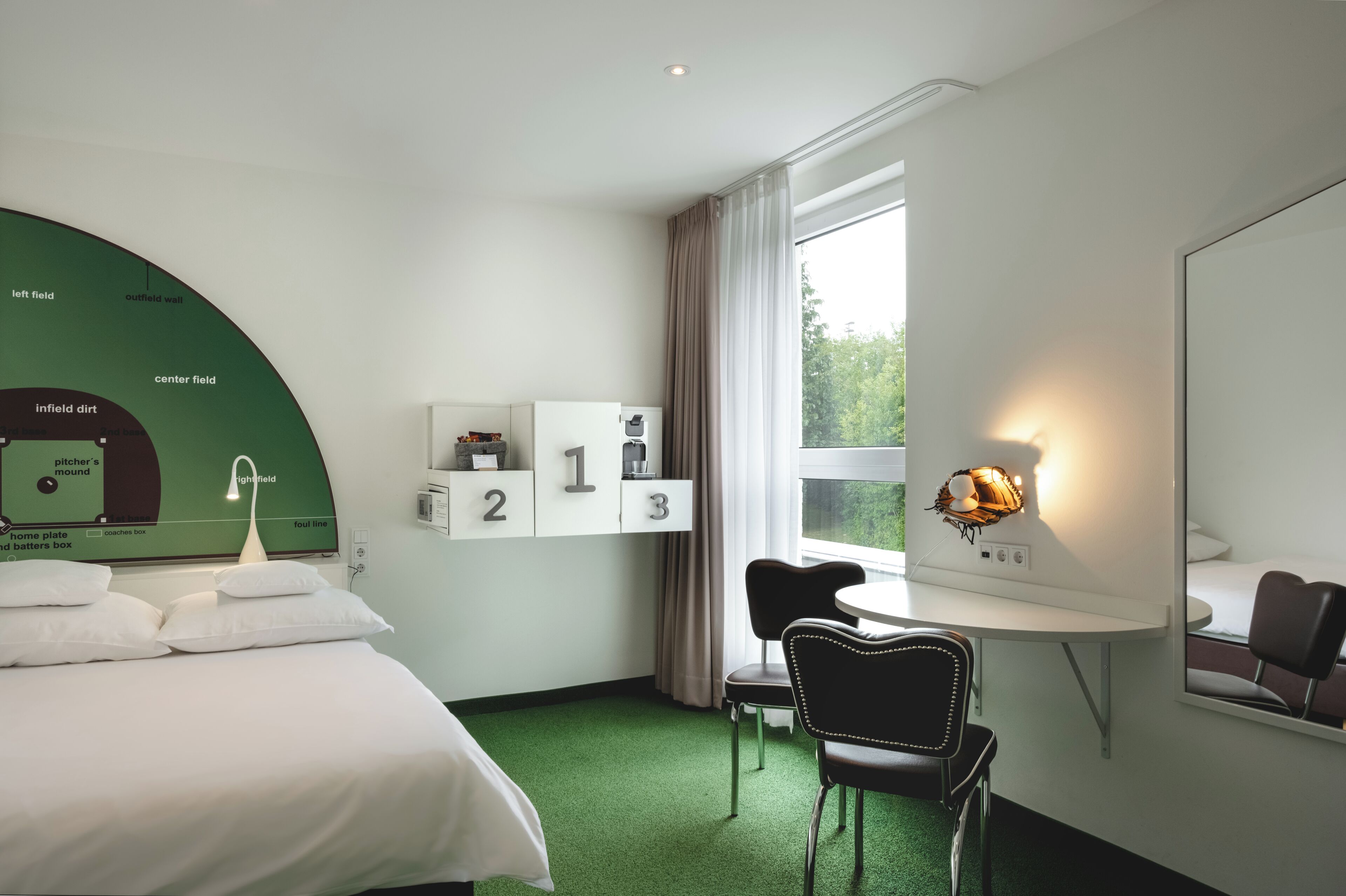 Habitación de hotel con cama, escritorio, silla, ventana con cortinas y un cartel con número en la pared.