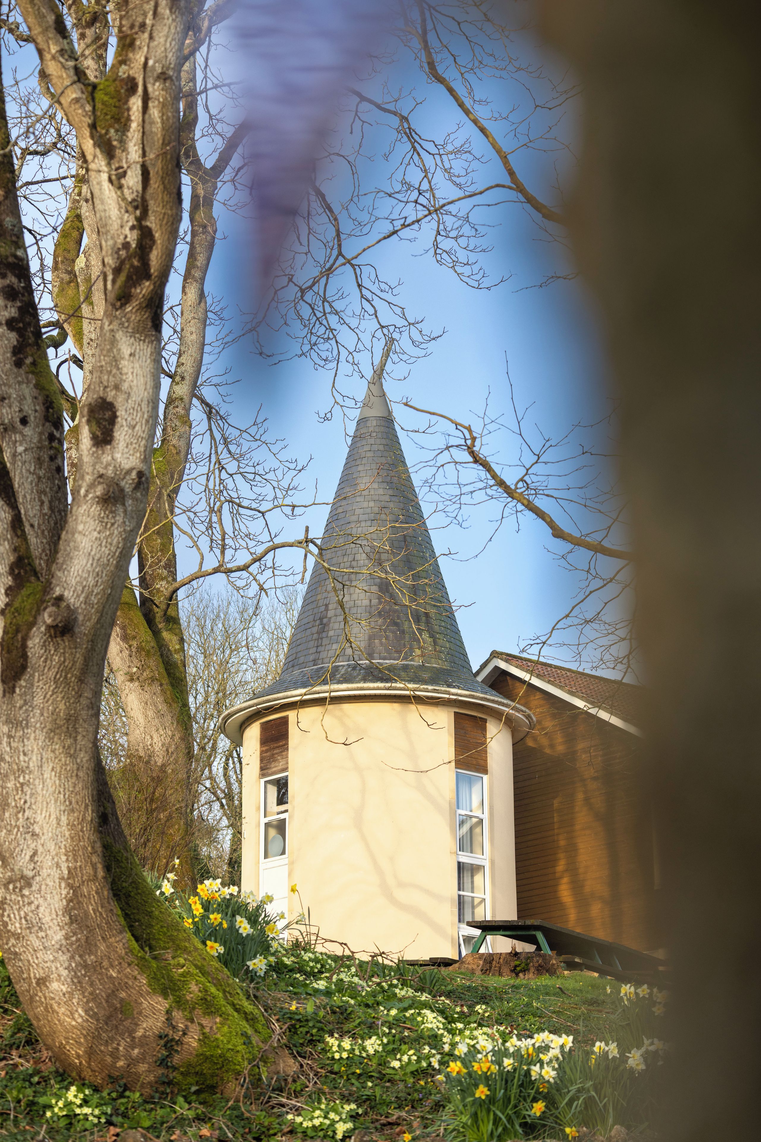 A building with a conical roof, surrounded by trees and flowering plants.