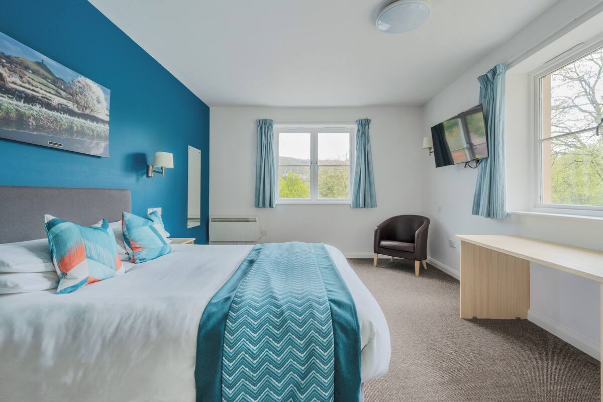 A bedroom with a bed, a desk, a chair, a TV, and a window with curtains.