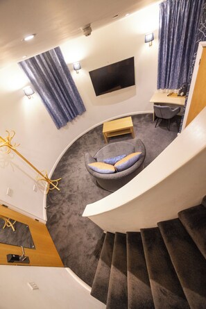 A modern interior with a curved staircase, a seating area with cushions, a flat-screen TV mounted on the wall, and a desk with a chair. - Wookey Hole Hotel (Wells)