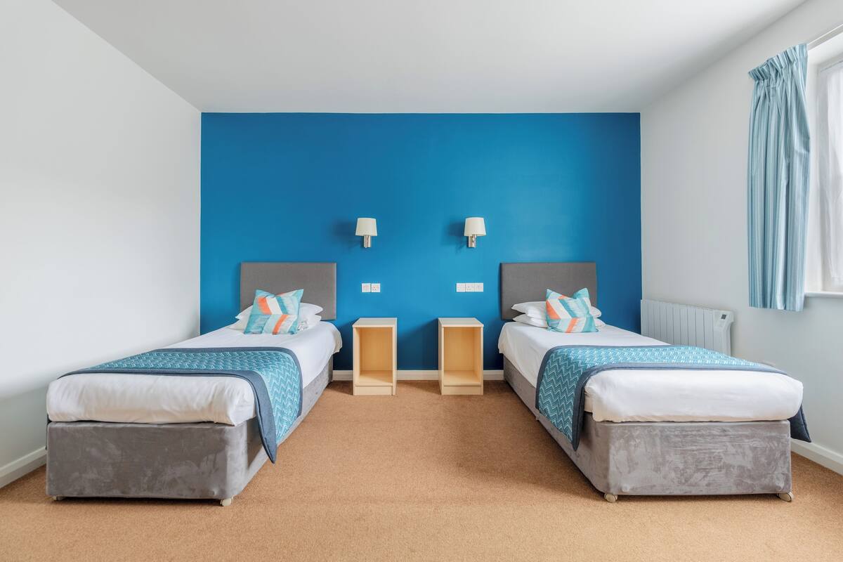 A twin room with two beds, a blue accent wall, bedside lamps, and a window with curtains.