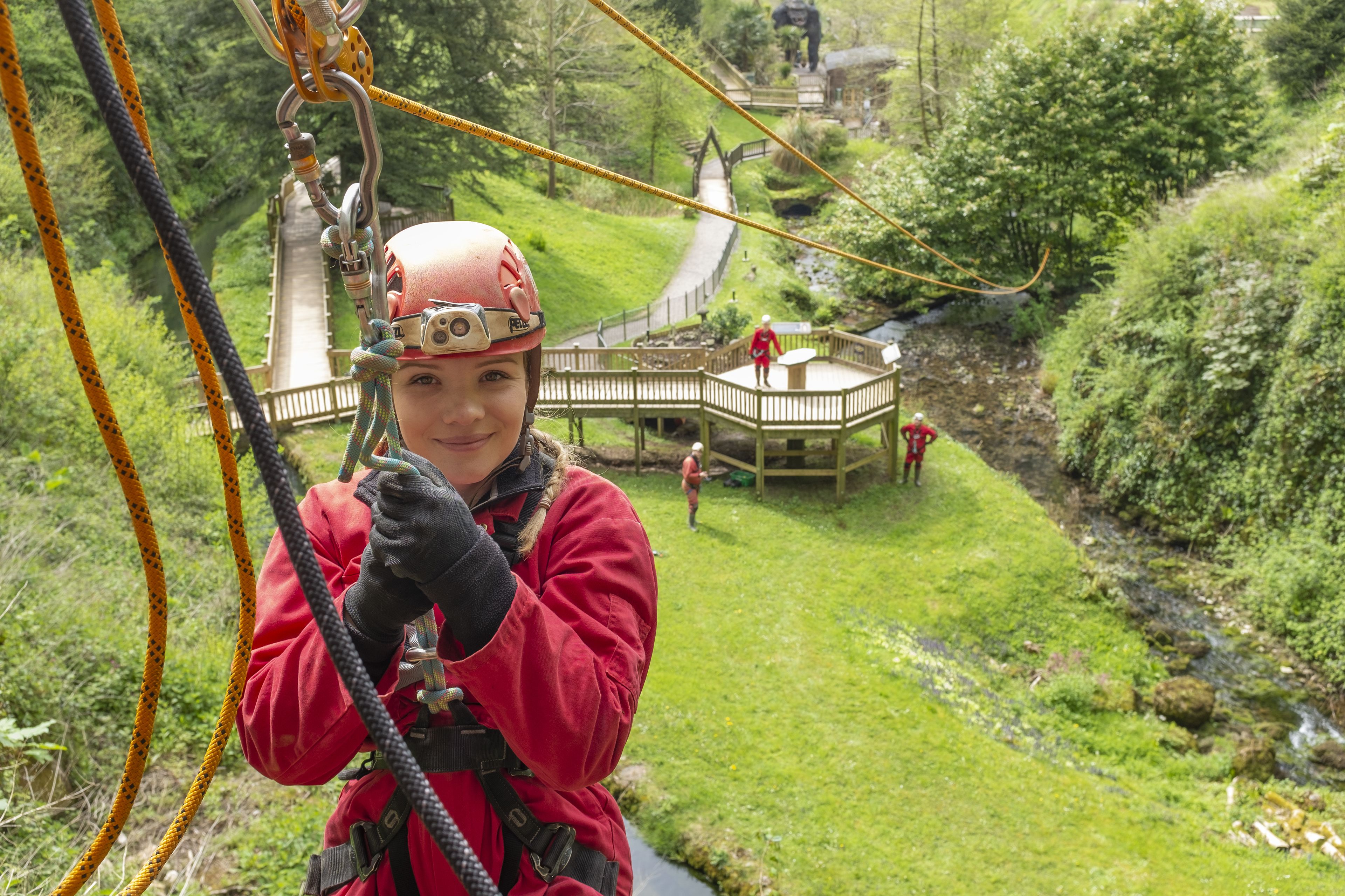 A person in a red jacket and helmet, holding onto a rope, with a wooden deck and greenery in the background.