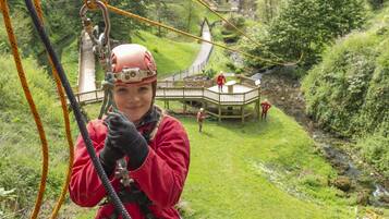 A person in a red jacket and helmet, holding onto a rope, with a wooden deck and greenery in the background.