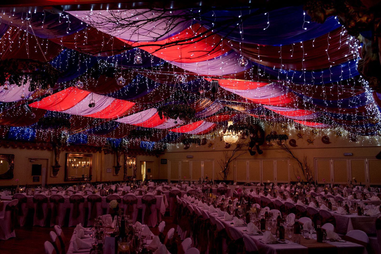 A banquet hall with tables set for a formal event, draped with red and white fabric and string lights.
