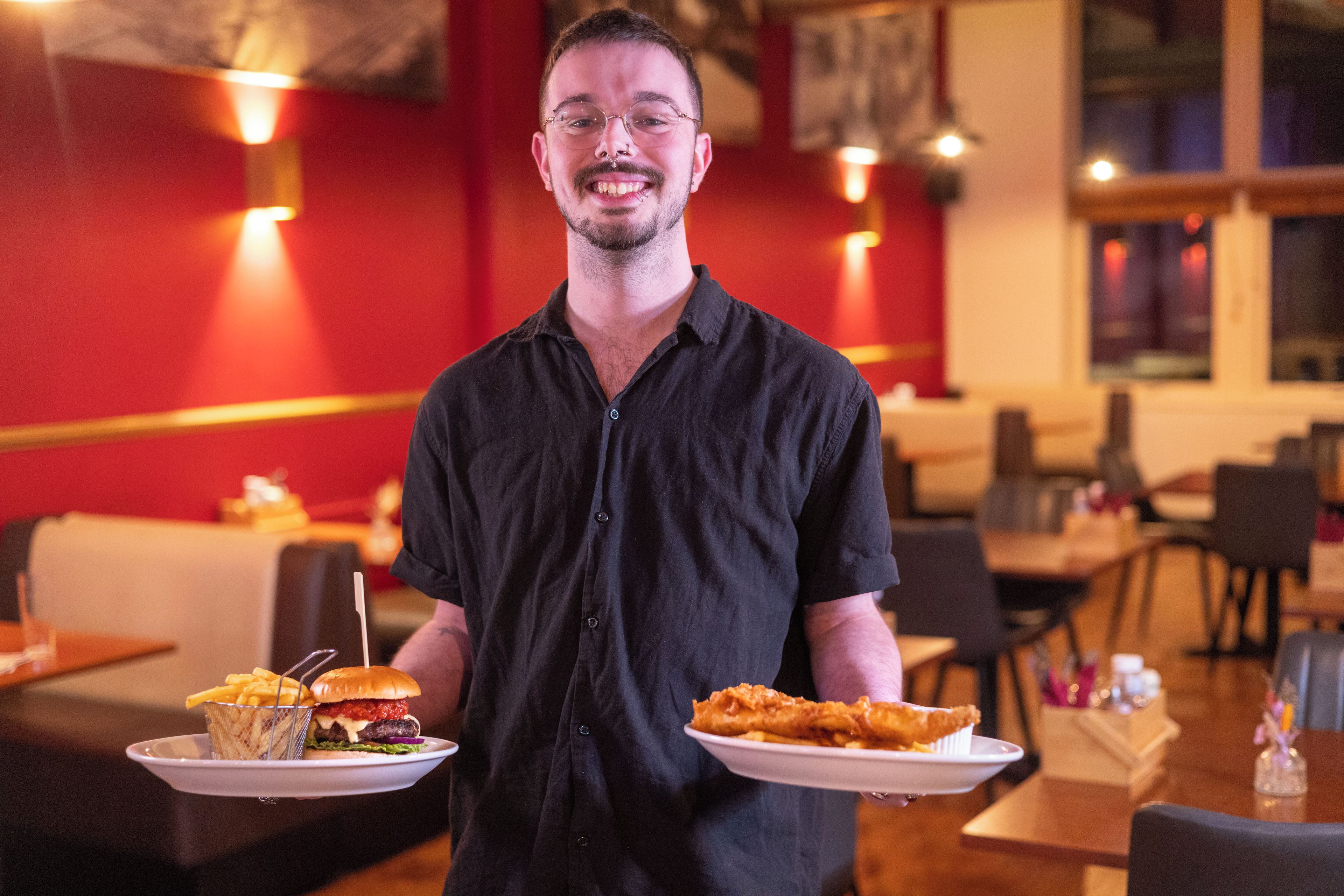 A person holding two plates of food in a restaurant with tables and chairs.