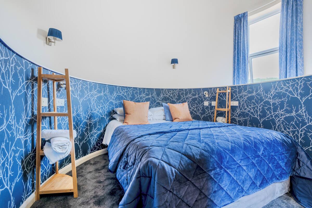 A bedroom with a curved ceiling, a bed with a blue quilt, a wooden ladder, and a wall-mounted shelf with towels.