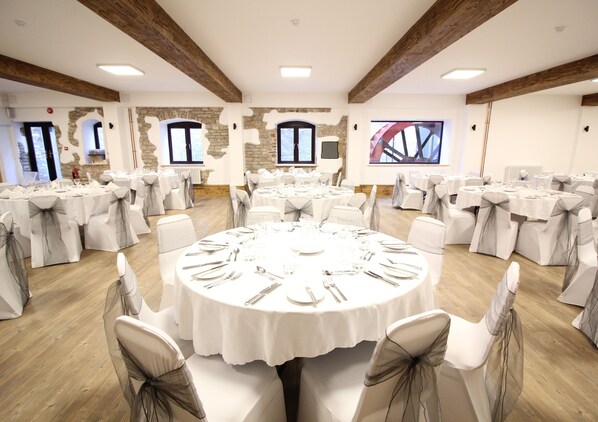 A banquet hall with round tables set for dining, white chairs with grey sashes, and exposed wooden beams on the ceiling. - Wookey Hole Hotel (Wells)
