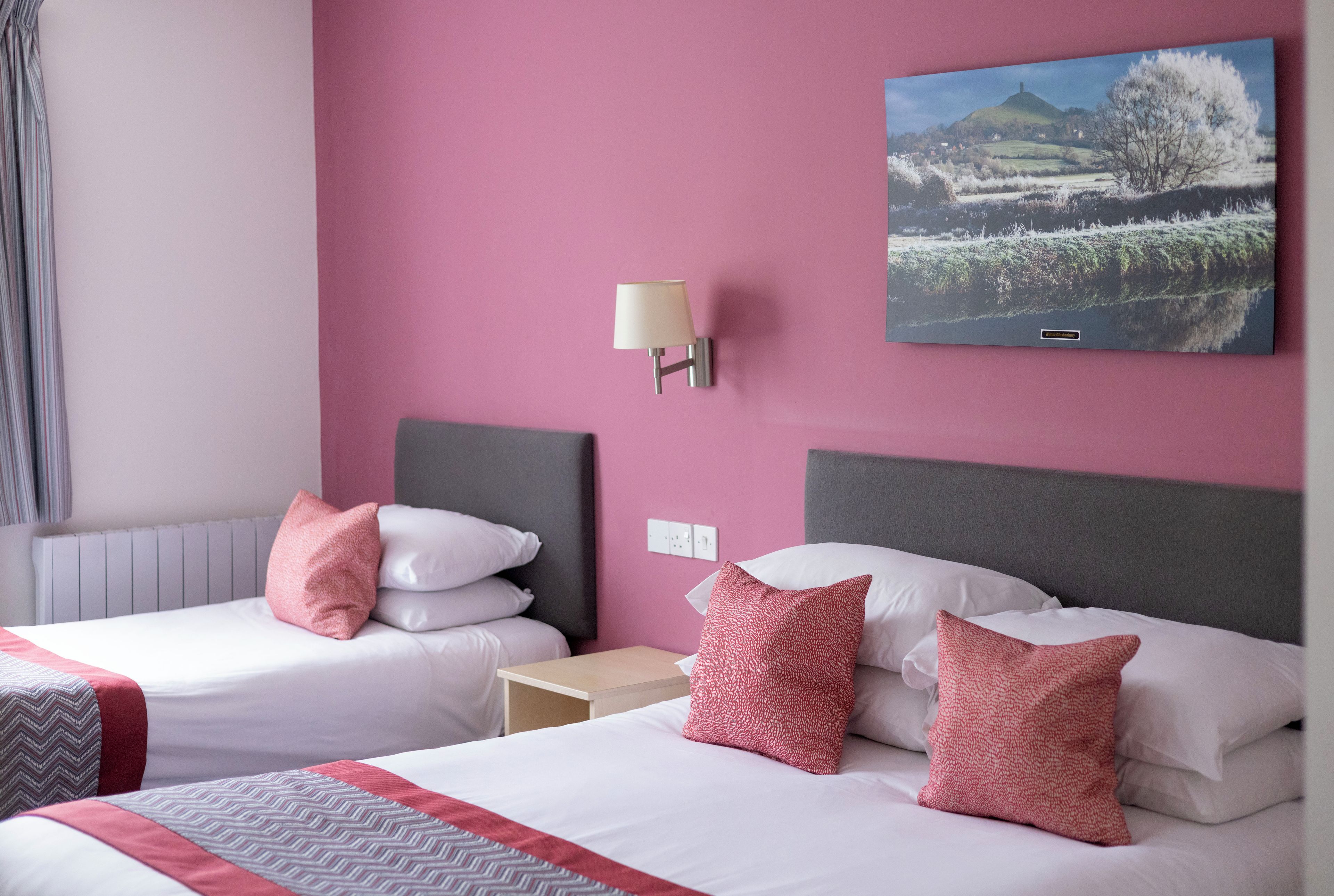 Une chambre d’hôtel avec deux lits, une table de chevet, une lampe fixée au mur et un tableau encadré.