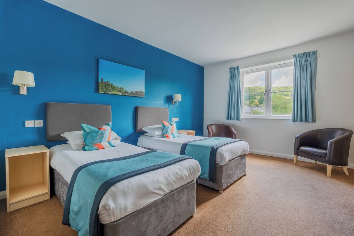 A hotel room with two beds, a window with curtains, a chair, and a painting on the wall.