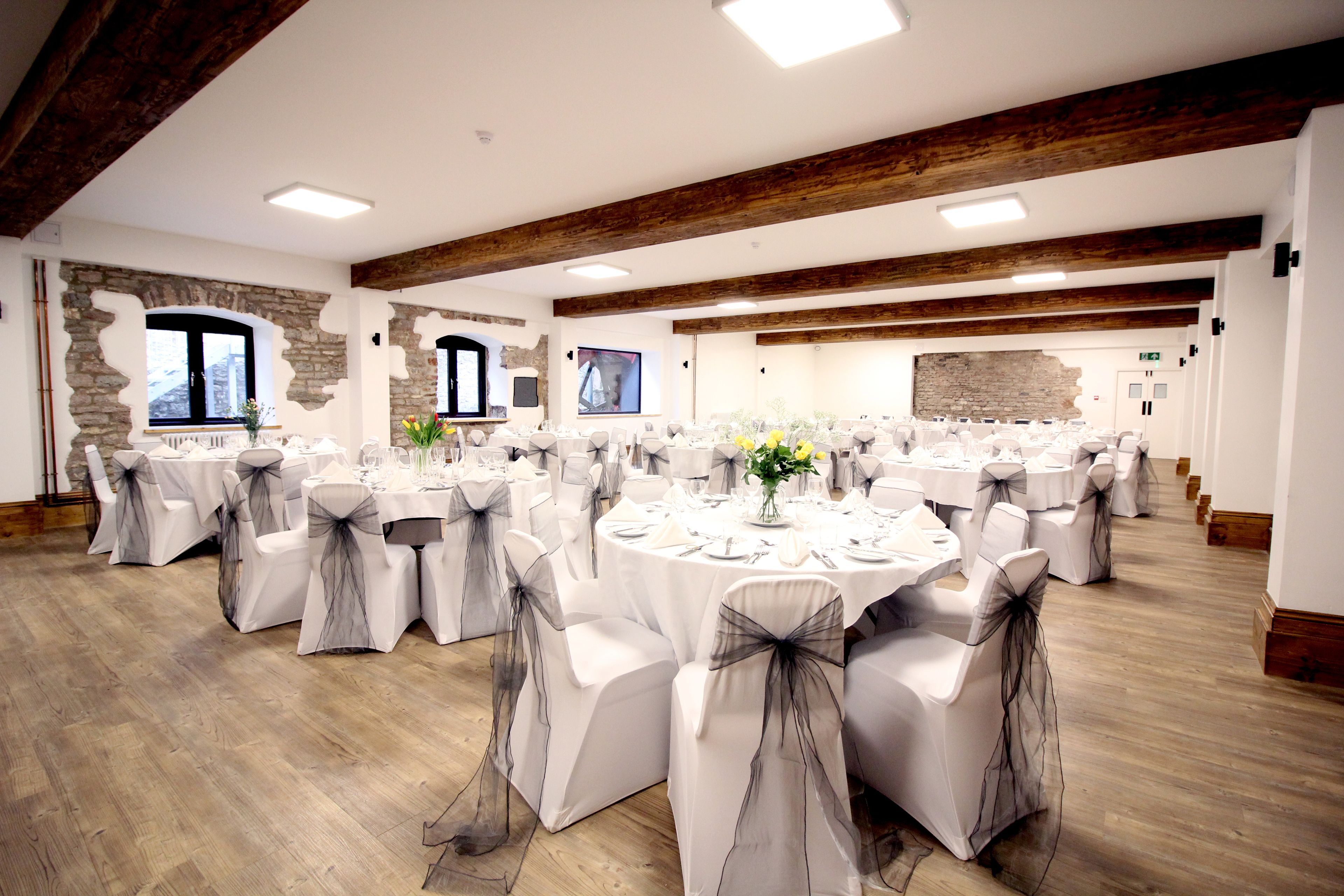 A banquet hall with round tables, white chairs with grey sashes, wooden beams on the ceiling, stone walls, and a large window.