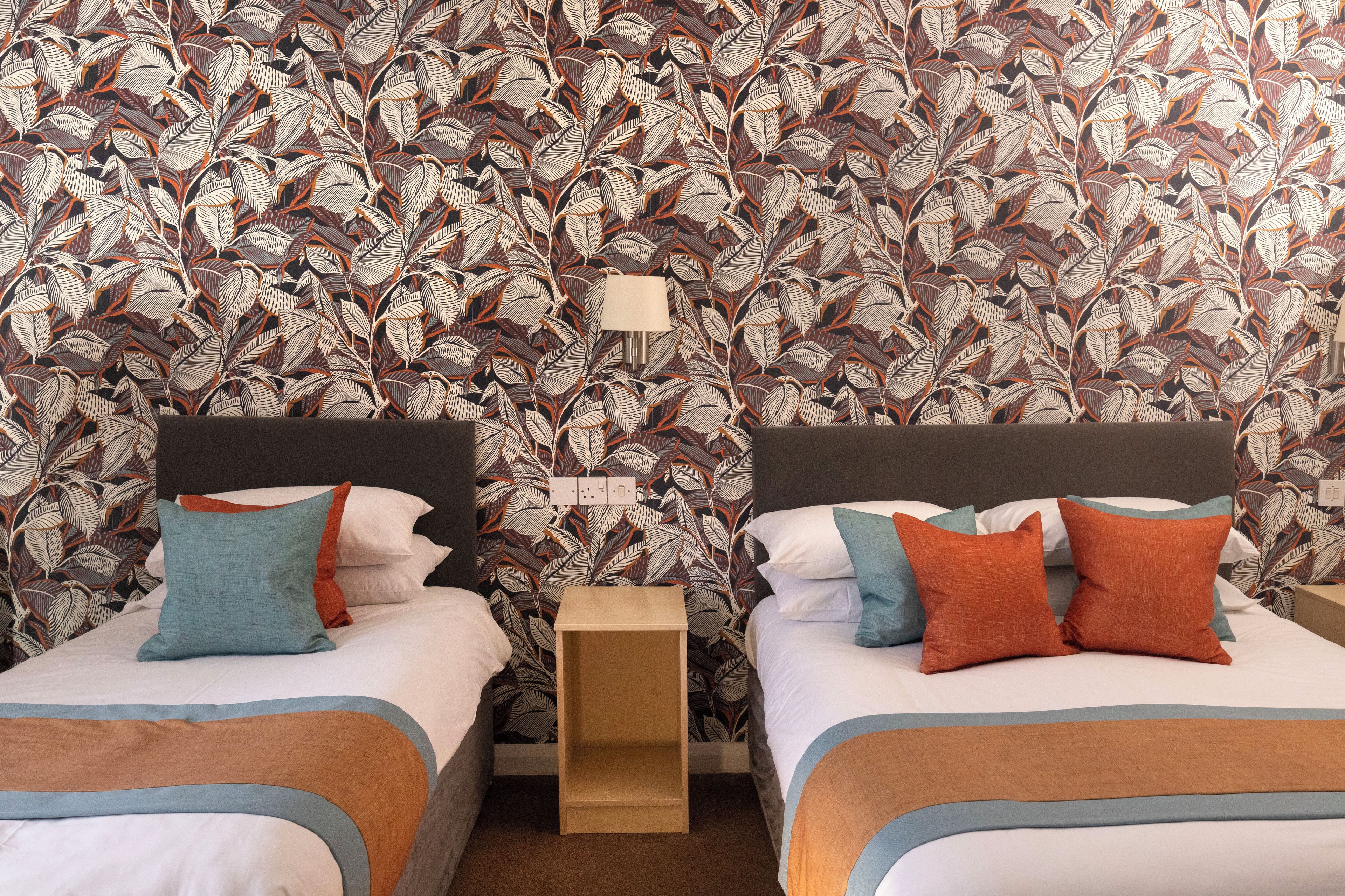 A hotel room with two beds, each with a headboard, white bedding, and multiple pillows in blue, orange, and teal.