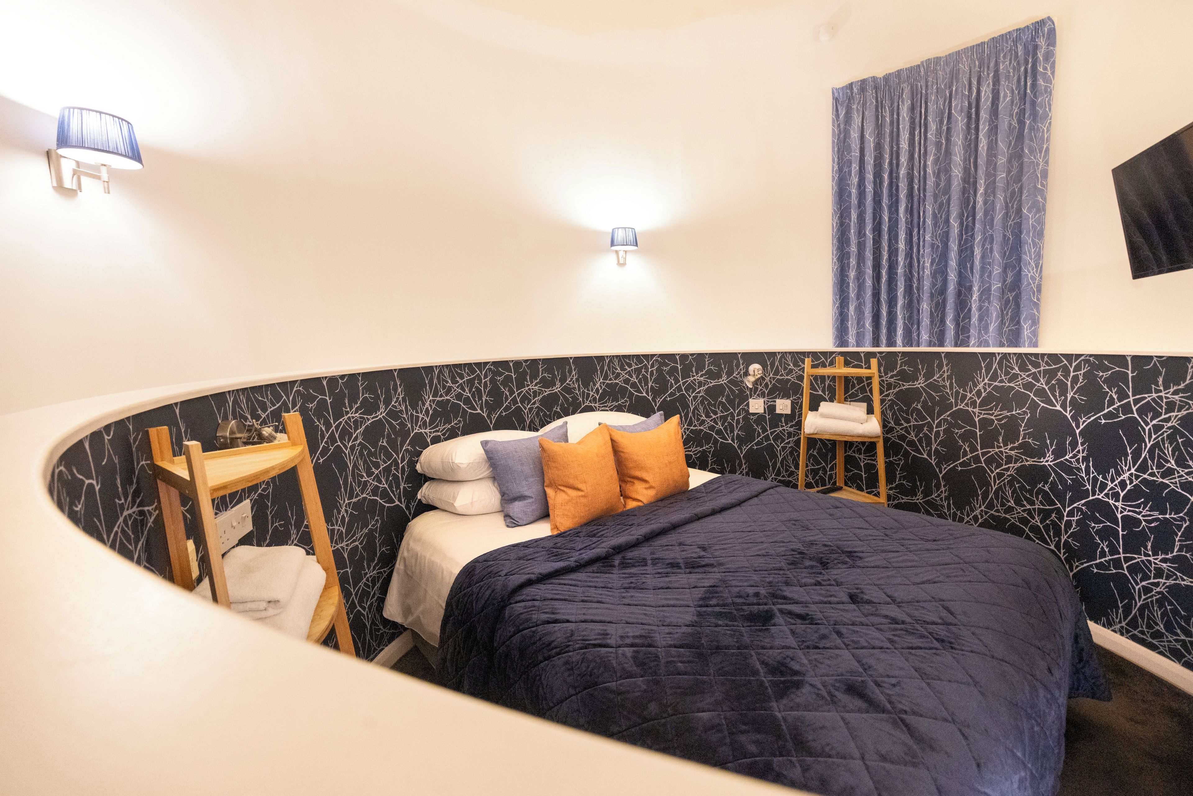 A bedroom with a curved ceiling, a bed with multiple pillows, a wooden ladder, and a wall-mounted shelf.